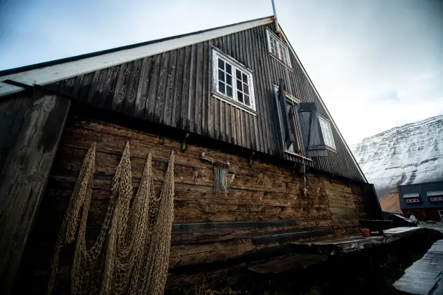 An old weathered wooden cabin with drying stockfish nets against a snow-dusted mountainside.