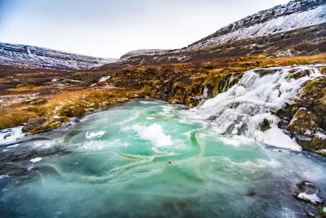 A turquoise glacial pool frozen over beneath an icy waterfall in a snowy valley, Iceland