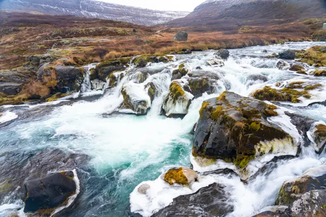 A turquoise river tumbling over rocky cascades through an autumnal highland valley.