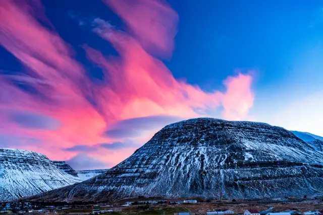 Vivid pink clouds drifting over snow-covered mountains above a small town, Iceland