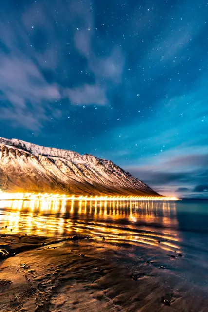 A line of golden town lights reflected on wet sand beneath a snowy mountain and starry sky, Iceland