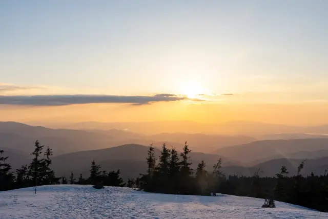 The sun setting over receding mountain ridges with snow and pines in the foreground