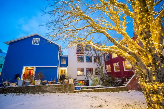 Brightly painted houses and a fairy-light-wrapped tree in fresh snow at blue hour.
